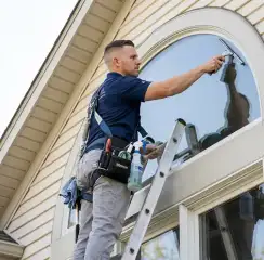 Professional window cleaner on a ladder using a squeegee to clean an exterior arched window, Savannah, GA.
